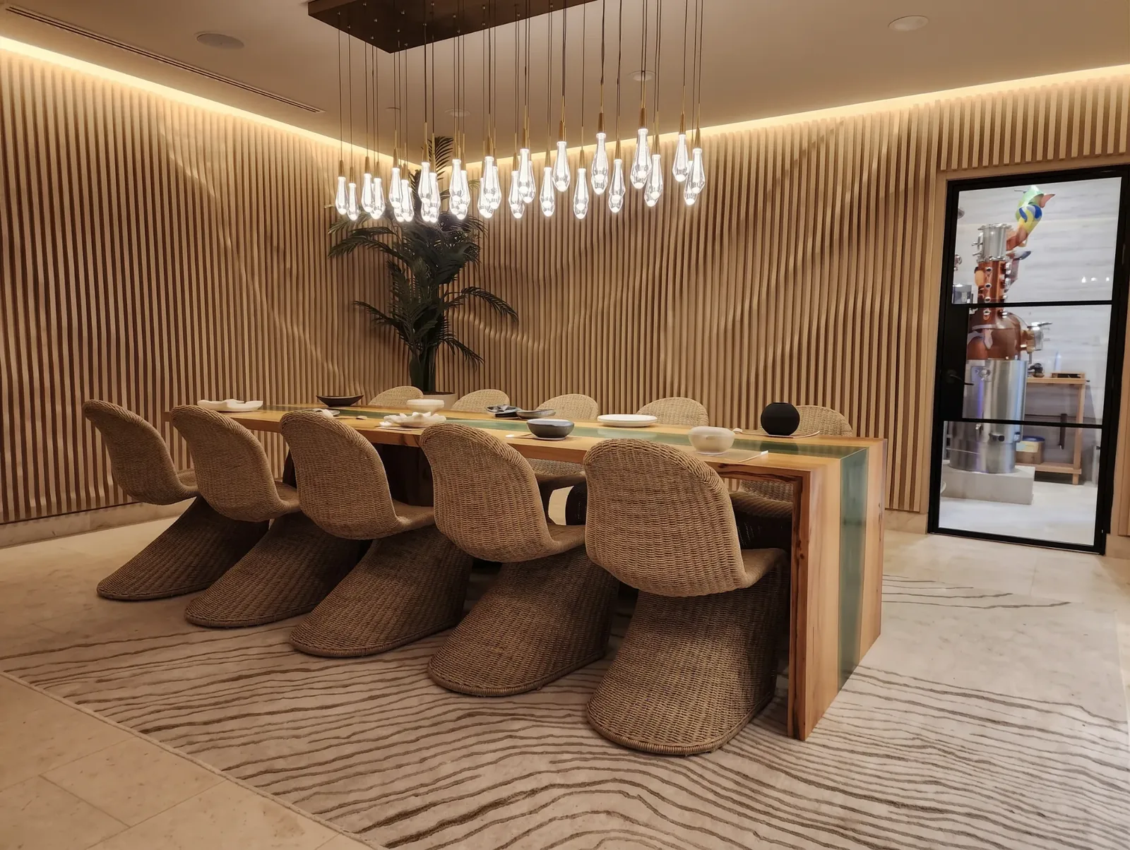 The private dining room at Mistura: a long wooden table with woven chairs under a cluster of glass pendant lights, with the copper still behind a glass door